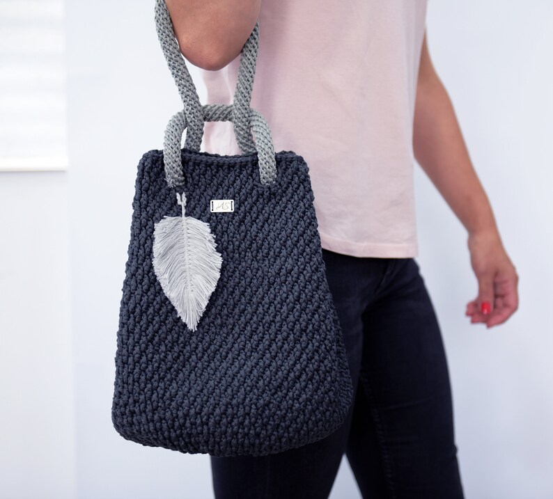 May include: A dark gray crocheted tote bag with a gray rope handle and a feather charm. The bag has a small silver tag with the letters "MC" on it.