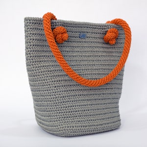 May include: A gray crocheted tote bag with orange rope handles. The handles have knots at the top.