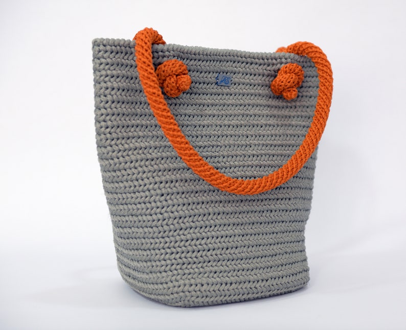 May include: A gray crocheted tote bag with orange rope handles. The handles have knots at the top.