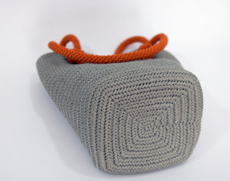 May include: A gray crocheted tote bag with orange handles. The bag has a square bottom and a woven texture.