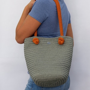 May include: A gray crocheted tote bag with orange handles and pom-poms. The bag has a small blue label with the letters "AS" on it.