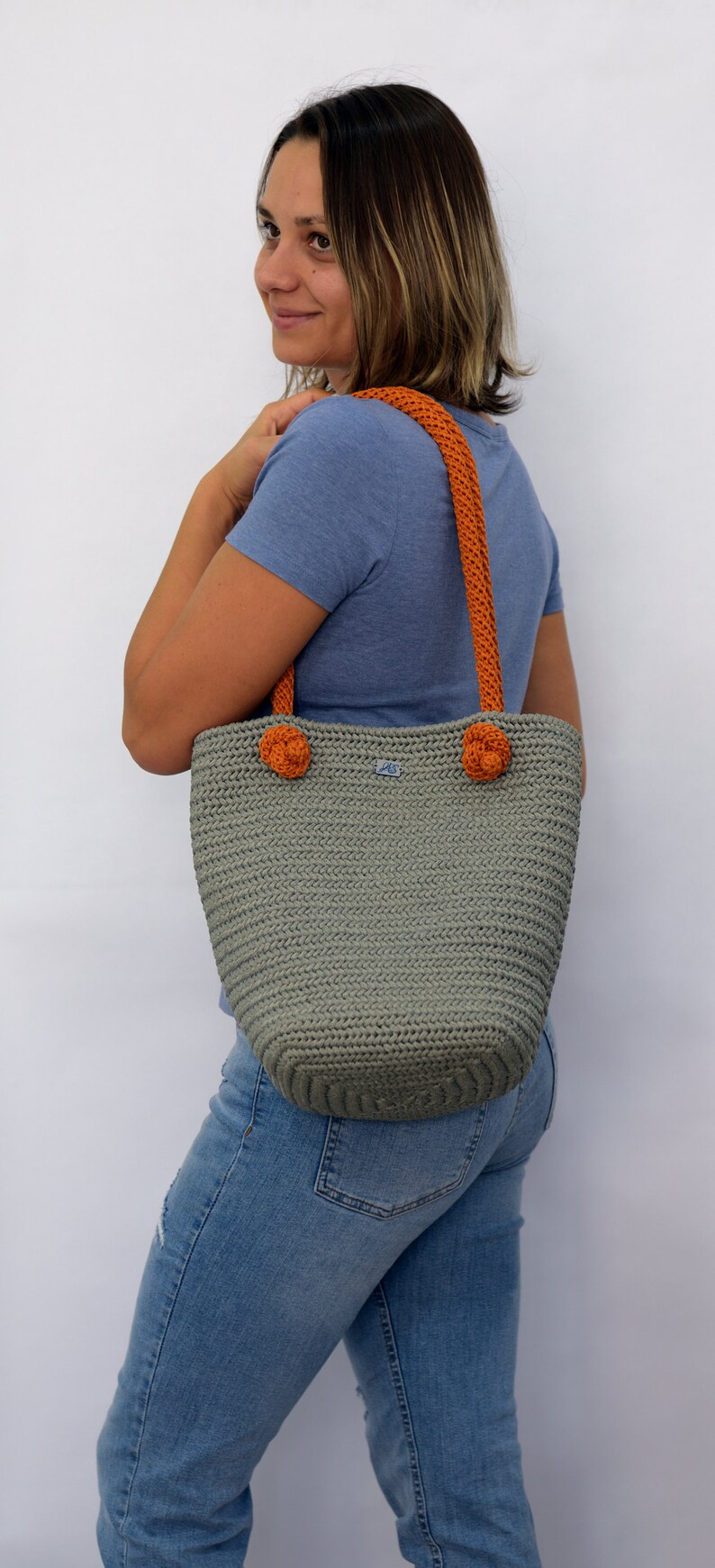 May include: A gray crocheted tote bag with orange handles and pom-poms. The bag has a small blue label with the letters "AS" on it.
