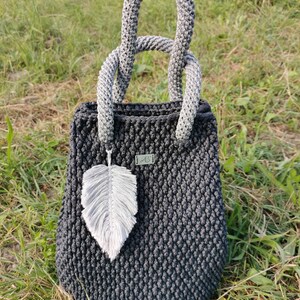 May include: A dark gray crocheted tote bag with a gray and white crocheted handle. The bag has a small white feather charm attached to it.