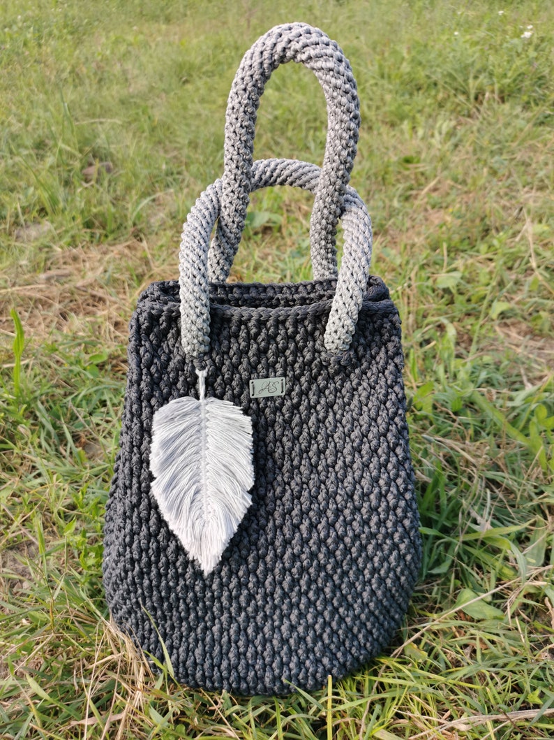 May include: A dark gray crocheted tote bag with a gray and white crocheted handle. The bag has a small white feather charm attached to it.
