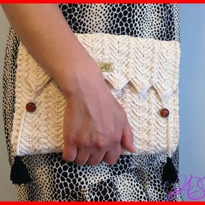 May include: Cream-colored crochet clutch with a chevron pattern, secured with two brown buttons and a flap closure. The clutch features black tassels and a small tag with initials. A stylish accessory.