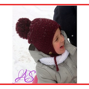 Könnte beinhalten: Ein Kind trägt eine burgunderfarbene Strickmütze mit Bommel und einen weißen Schal. Die Mütze hat eine Klappe, die die Ohren bedeckt und unter dem Kinn gebunden wird.