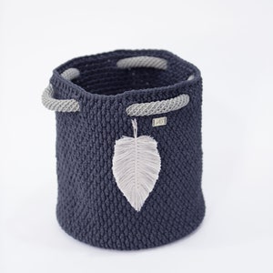 May include: A dark blue crocheted storage basket with gray rope handles and a white feather accent. The basket is made of a thick, textured yarn.