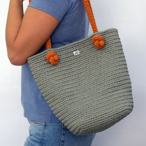 May include: A gray crocheted tote bag with orange straps and a small label that says "JAS". The bag has two orange crocheted flower accents on the front.