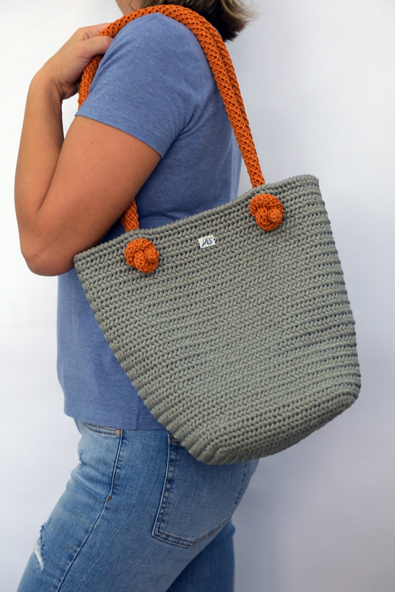 May include: A gray crocheted tote bag with orange straps and a small label that says "JAS". The bag has two orange crocheted flower accents on the front.