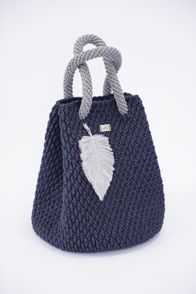 May include: A dark blue crocheted tote bag with a gray crocheted handle and a white feather charm. The bag has a small silver tag with the letters "AS" on it.