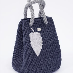 May include: A dark blue crocheted tote bag with a gray crocheted handle and a white feather charm. The bag has a small silver tag with the letters "AS" on it.