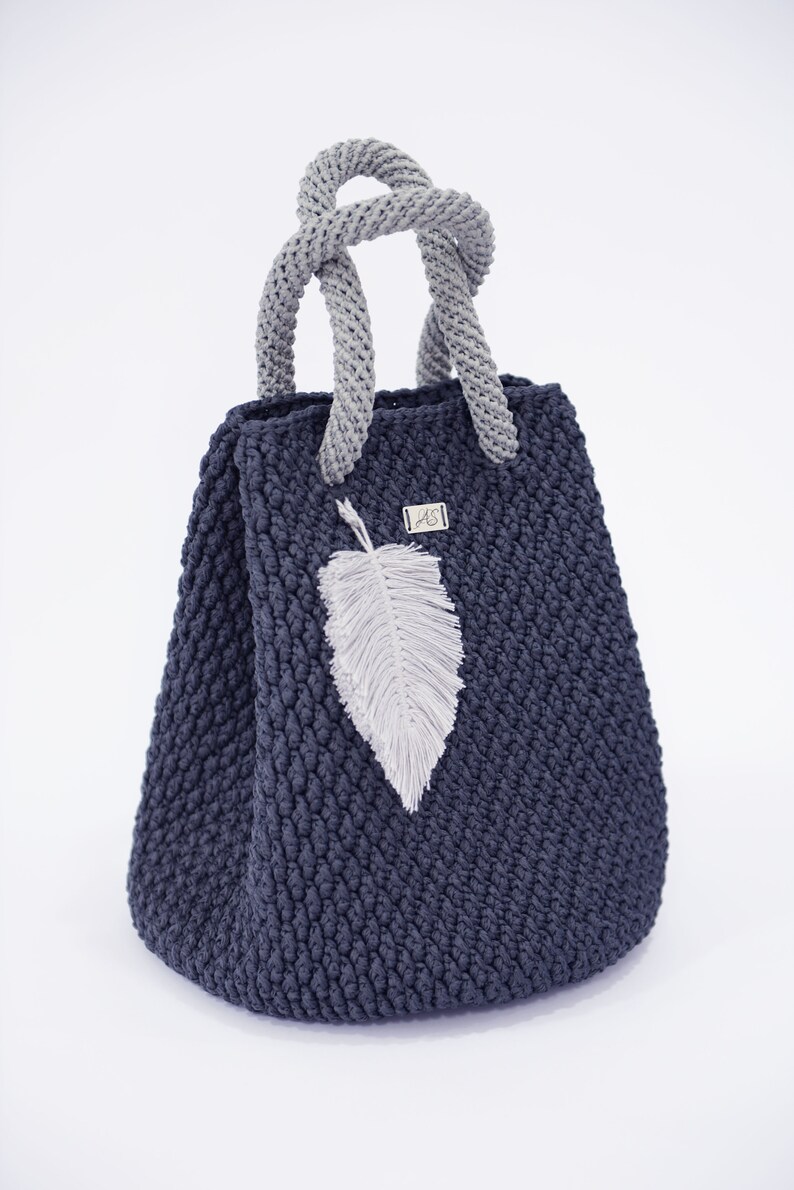 May include: A dark blue crocheted tote bag with a gray crocheted handle and a white feather charm. The bag has a small silver tag with the letters "AS" on it.