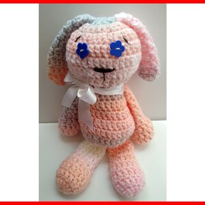 May include: A crocheted stuffed animal in the shape of a dog with pink, gray, and white yarn. The dog has blue flower-shaped buttons for eyes and a white ribbon around its neck.