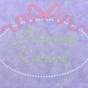 Peut inclure: Un cadre ovale violet et blanc avec un nœud de ruban rose et des accents de perles. Le texte "Ariana Grace" est brodé en vert à l'intérieur du cadre.