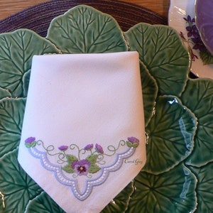 May include: A white linen napkin with an embroidered design of purple flowers and green leaves. The napkin is on a green and purple ceramic plate with a leafy design.