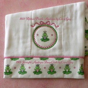 May include: White burp cloth with a pink border and a green frog applique with a crown. The cloth has the text "1901 Straight Applique Burp Cloth by Carol Guy" printed on it.