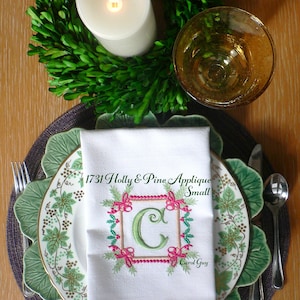Peut inclure: Une serviette blanche avec un motif brodé vert, rouge et rose représentant une lettre C et le texte "1731 Holly & Pine Applique Small Carol Guy". La serviette est posée sur une assiette à motifs verts et blancs avec un bord brun.