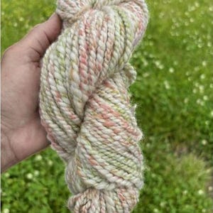May include: A skein of hand-spun yarn in shades of cream, pale green, and pink. The thick yarn is held in a hand, with the background showing green grass and small white flowers. The yarn appears soft and textured, suitable for knitting or crochet projects.