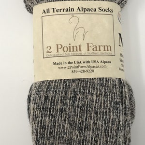 Puede incluir: Un par de calcetines de alpaca jaspeados en marr&oacute;n claro y negro envueltos en una banda de papel con la etiqueta All Terrain Alpaca Socks, 2 Point Farm, Distinguished Suri Alpacas of Northern Kentucky, Made in the USA with USA Alpaca.