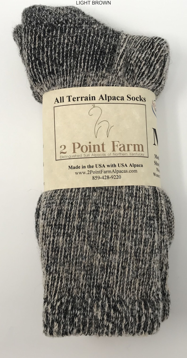 Puede incluir: Un par de calcetines de alpaca jaspeados en marr&oacute;n claro y negro envueltos en una banda de papel con la etiqueta All Terrain Alpaca Socks, 2 Point Farm, Distinguished Suri Alpacas of Northern Kentucky, Made in the USA with USA Alpaca.