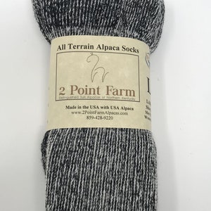 Puede incluir: Un par de calcetines de alpaca jaspeados en gris oscuro y blanco envueltos en una etiqueta de papel color canela. La etiqueta dice All Terrain Alpaca Socks, 2 Point Farm, Distinguished Suri Alpacas of Northern Kentucky, Made in the USA.
