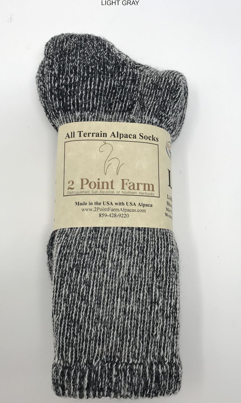 Puede incluir: Un par de calcetines de alpaca jaspeados en gris oscuro y blanco envueltos en una etiqueta de papel color canela. La etiqueta dice All Terrain Alpaca Socks, 2 Point Farm, Distinguished Suri Alpacas of Northern Kentucky, Made in the USA.