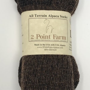 Puede incluir: Un par de calcetines de alpaca de color marr&oacute;n oscuro envueltos en una banda de papel beige etiquetada como All Terrain Alpaca Socks, 2 Point Farm, Made in the USA with USA Alpaca, con el sitio web www.2PointFarmAlpacas.com y el tel&eacute;fono 859-428-9220.