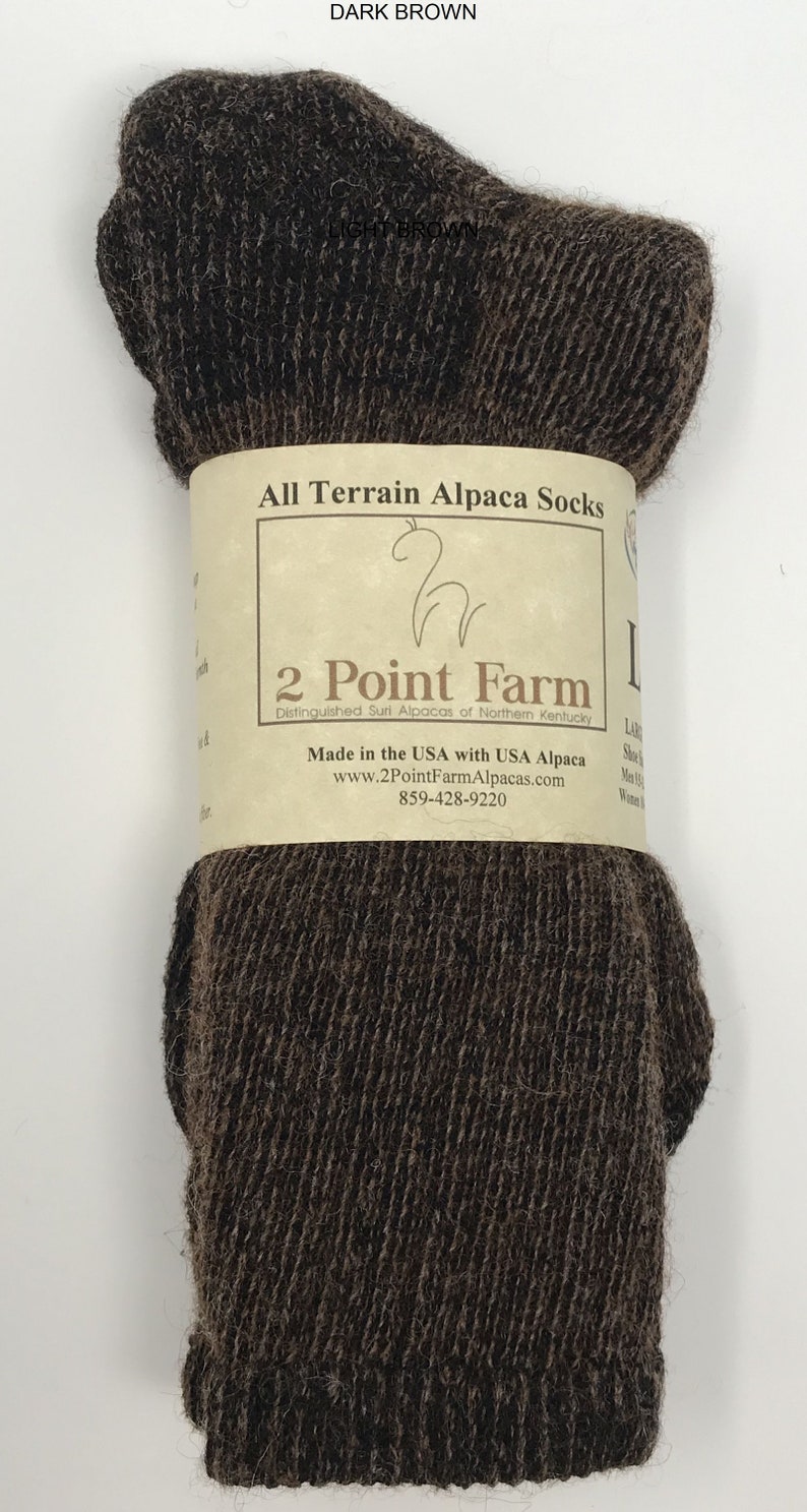 Puede incluir: Un par de calcetines de alpaca de color marr&oacute;n oscuro envueltos en una banda de papel beige etiquetada como All Terrain Alpaca Socks, 2 Point Farm, Made in the USA with USA Alpaca, con el sitio web www.2PointFarmAlpacas.com y el tel&eacute;fono 859-428-9220.