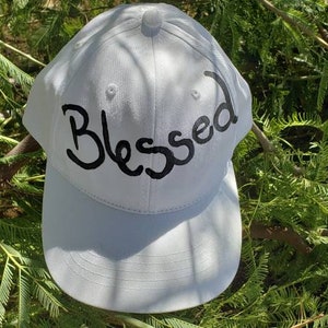 May include: A white baseball cap with the word "Blessed" written in black cursive lettering.