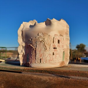 May include: A cylindrical ceramic vessel with a ruffled, uneven top edge. The exterior features a textured, relief-style depiction of a multi-story stone structure in shades of tan, pink, and brown, with small rectangular openings and green accents.