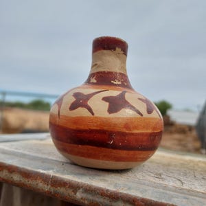 May include: A small, handcrafted ceramic vase with a rounded body and a narrow neck. The vase features horizontal bands of brown and tan, with brown star-like shapes. The vase is approximately 5 inches tall.