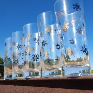 May include: A set of five clear glass tumblers arranged in a row, each decorated with a repeating pattern of metallic gold and black snowflake designs. The tall, cylindrical drinking glasses feature a smooth finish and a thick, weighted base.