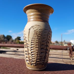 May include: A cylindrical ceramic vase with a flared rim, featuring a light tan matte finish and a dark brown glazed neck. The body is decorated with deeply carved, vertical fern leaf patterns that wrap around the exterior of the vessel.