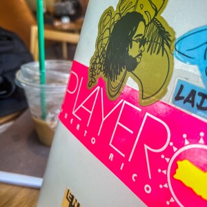 A sticker on a laptop of a Black man with locs in front of tropical plants on a yellow green background.