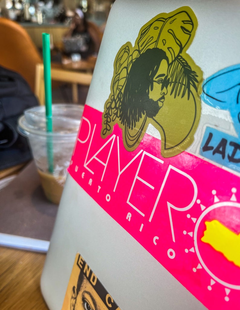 A sticker on a laptop of a Black man with locs in front of tropical plants on a yellow green background.