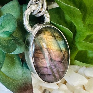 May include: A silver pendant necklace with an oval labradorite gemstone. The stone has iridescent colours of purple, gold, and green. The pendant hangs from a silver chain, with a unique, textured design.