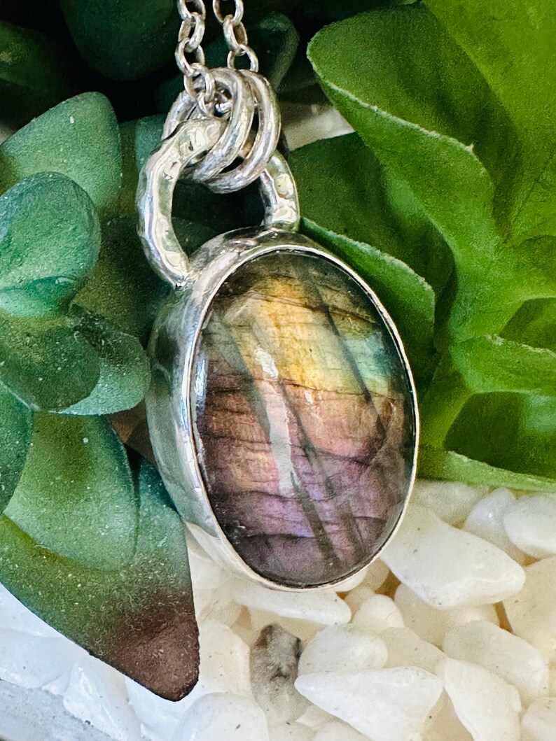 May include: A silver pendant necklace with an oval labradorite gemstone. The stone has iridescent colours of purple, gold, and green. The pendant hangs from a silver chain, with a unique, textured design.