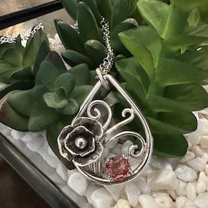 May include: A silver pendant necklace with a delicate floral design and a pink gemstone. The pendant is shaped like a teardrop and features a stylized flower with a small pearl in the center. The necklace is on a silver chain.