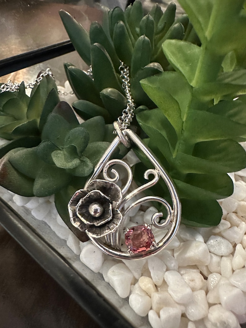 May include: A silver pendant necklace with a delicate floral design and a pink gemstone. The pendant is shaped like a teardrop and features a stylized flower with a small pearl in the center. The necklace is on a silver chain.