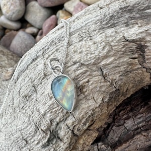 May include: A silver necklace with a pear-shaped pendant. The pendant has a rainbow-colored, iridescent stone set in a silver bezel. The necklace is displayed on a piece of weathered wood.