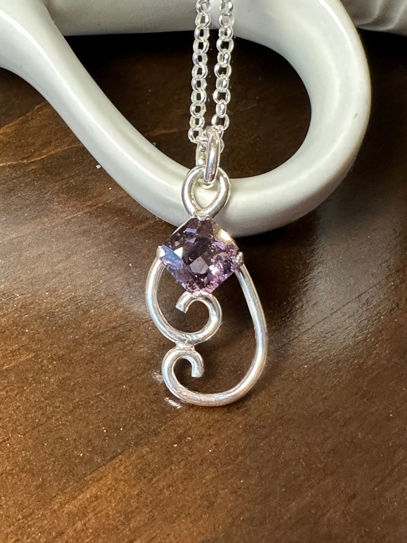 May include: A silver necklace with a delicate pendant featuring a purple gemstone and a swirling design.