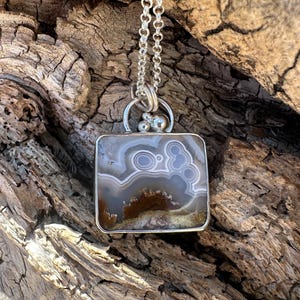 May include: A silver necklace featuring a square pendant with a natural stone. The stone displays a pattern of gray, white, and brown swirls and circles. The pendant is suspended from a delicate silver chain, creating an elegant accessory.