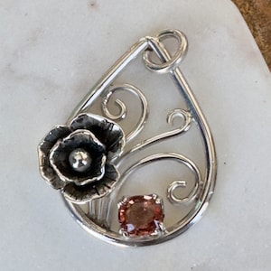 May include: A silver pendant with a delicate floral design and a pink gemstone. The pendant is shaped like a teardrop and features a stylized flower with a small pearl in the center.
