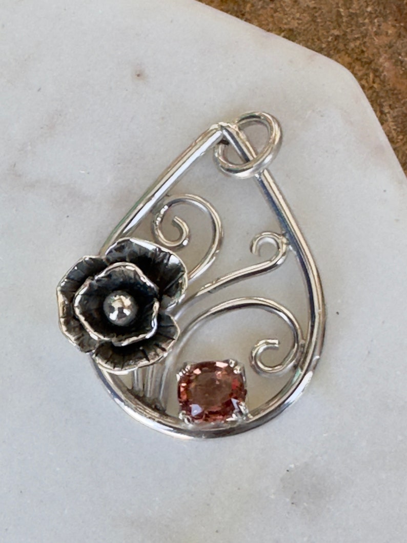 May include: A silver pendant with a delicate floral design and a pink gemstone. The pendant is shaped like a teardrop and features a stylized flower with a small pearl in the center.
