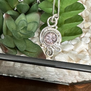 May include: A silver necklace with a delicate pendant featuring a pink gemstone surrounded by a silver filigree design. The pendant is resting on a bed of white pebbles.