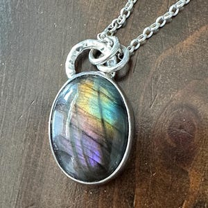 May include: A silver necklace with an oval pendant showcasing an iridescent gemstone. The stone exhibits a play of colours, including purple, blue, and yellow. The pendant hangs from a fine silver chain, creating an elegant piece of jewellery.
