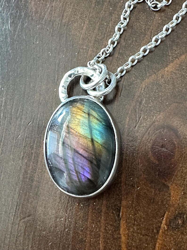 May include: A silver necklace with an oval pendant showcasing an iridescent gemstone. The stone exhibits a play of colours, including purple, blue, and yellow. The pendant hangs from a fine silver chain, creating an elegant piece of jewellery.
