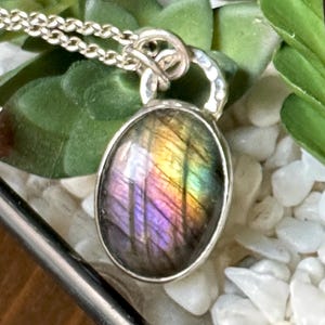 May include: Silver necklace featuring an oval labradorite pendant. The pendant showcases a shimmering display of colours, including purple, yellow, and green. The necklace is set against a backdrop of green foliage and white pebbles.