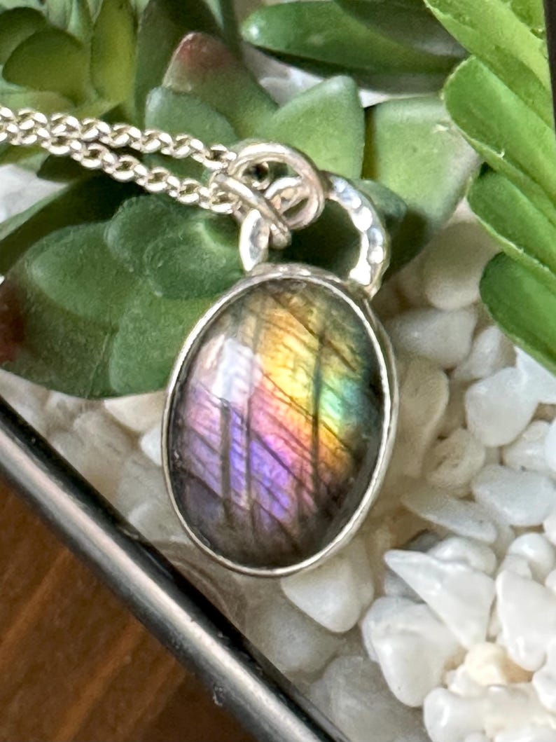 May include: Silver necklace featuring an oval labradorite pendant. The pendant showcases a shimmering display of colours, including purple, yellow, and green. The necklace is set against a backdrop of green foliage and white pebbles.