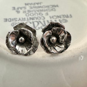 May include: A pair of silver flower-shaped earrings with a textured surface. The earrings are on a white background.